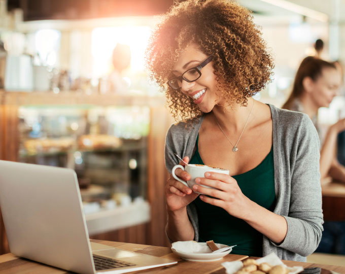 A woman in a cafe shop holding a mug and looking at a laptop.