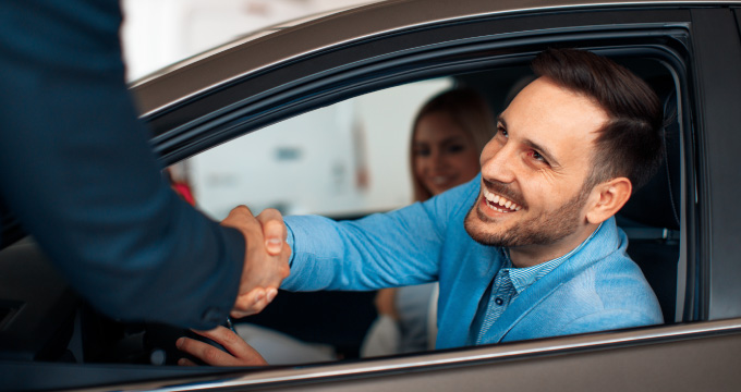 Man sitting in a new car shaking the hand of the salesman. 
