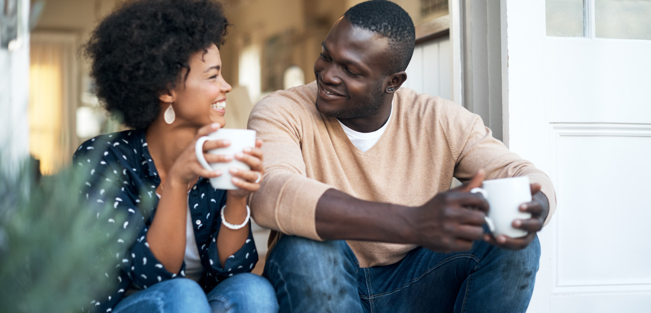 Couple sitting on steps outside their home holding mugs and looking at each other.